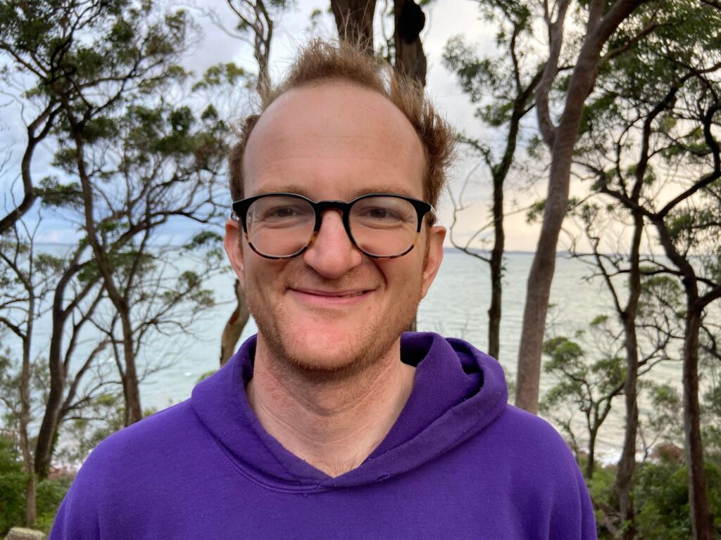 A photo of a man with red hair and glasses smiling in front of some trees.