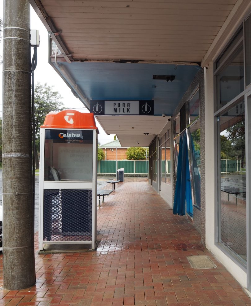A Telstra public payphone booth opposite a milkbar on a suburban strip