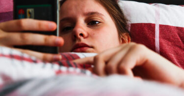 A girl lies in bed with a glum face looking at her phone.
