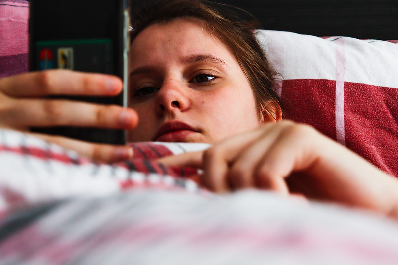 A girl lies in bed with a glum face looking at her phone.